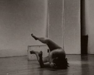 A nude woman with her legs raised and arms bent falls on the floor on her side.
