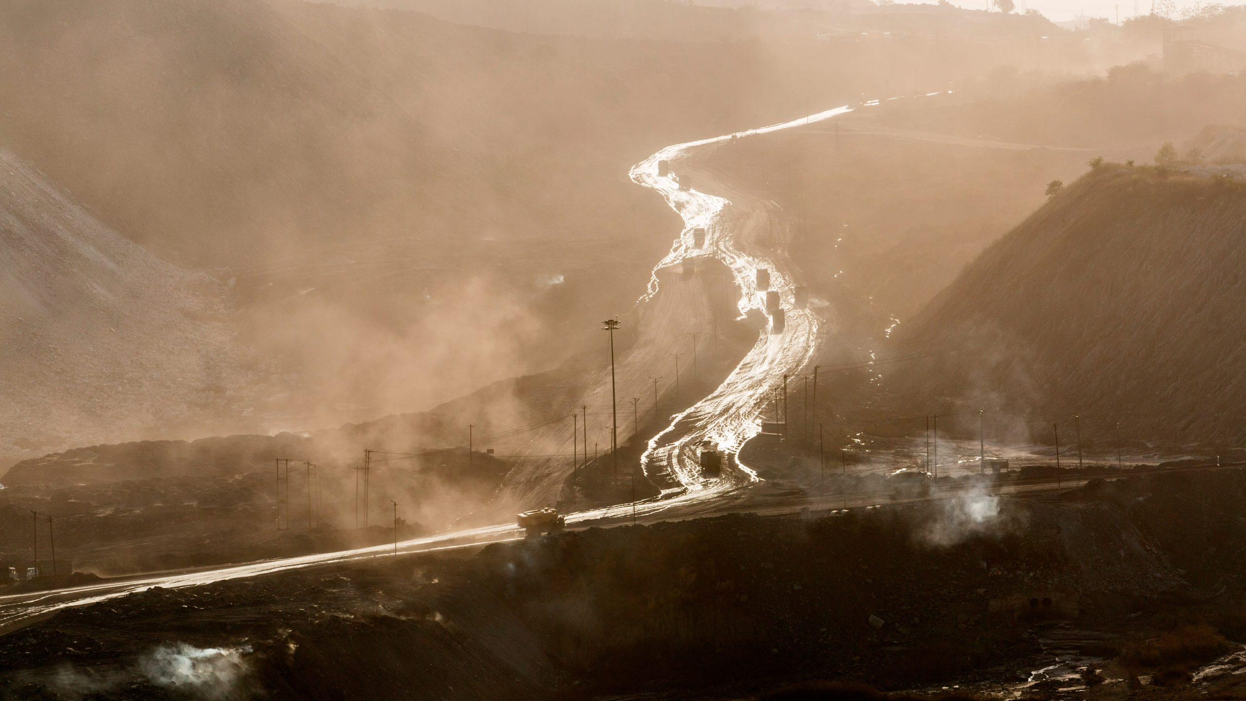 A landscape shot of mountains with winding roads, and trucks carrying mined material. The road seemingly glows under the sun which is shrouded by smoke clouds